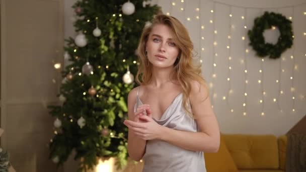 Porträtt av en vacker ung kvinna i en festlig silveraftonsklänning med öppna axlar. Julafton och nyårsafton. Julgran med kransar och ljus i bakgrunden. — Stockvideo
