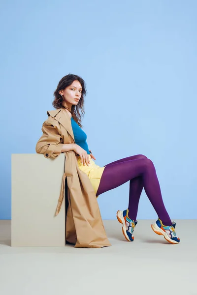 Bej rengi siperli, sarı kısa etekli, mor taytlı, mavi ve küblü güneş gözlüklü genç bir moda kızı..