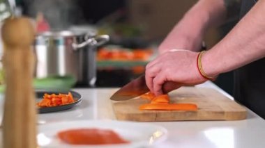 Bir erkek şef, restoranın mutfağında sebze salatası hazırlayan önlüklü bir profesyoneldir. Aşçı kesme tahtasındaki bir bıçakla çiğ havucu keser..
