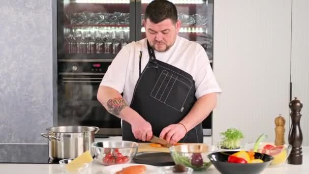Bir erkek şef, restoranın mutfağında sebze salatası hazırlayan önlüklü bir profesyoneldir. Aşçı kesme tahtasındaki bir bıçakla çiğ havucu keser.. — Stok video