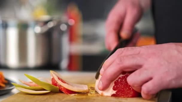 De mannelijke chef-kok reinigt en snijdt grapefruit op een houten snijplank in de keuken van de restaurants. Koken. — Stockvideo