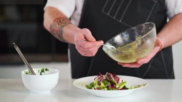 De mannelijke chef vult de salade met saus en boter. De kok voegt de saus toe met een lepel uit een glazen kom. Koken in de keuken van het restaurant. — Stockvideo