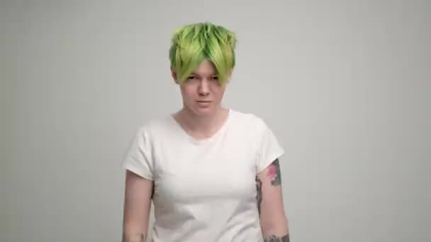 A young girl with a short pixie haircut and green hair in a white T-shirt on a light background. A woman poses in the studio, smiles and shows emotions. — Stock Video