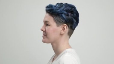 Kısa saçlı, koyu mavi saçlı, beyaz tişörtlü, açık arkaplanda genç bir kız. Bir kadın stüdyoda poz veriyor, gülümsüyor ve duygularını gösteriyor..