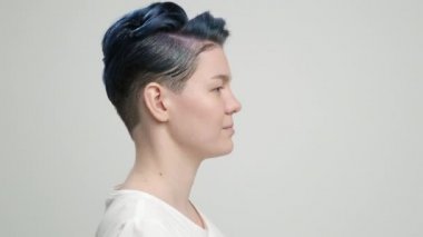 Kısa saçlı, koyu mavi saçlı, beyaz tişörtlü, açık arkaplanda genç bir kız. Bir kadın stüdyoda poz veriyor, gülümsüyor ve duygularını gösteriyor..