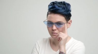 Kısa saçlı, koyu mavi saçlı, beyaz tişörtlü, açık arkaplanda genç bir kız. Bir kadın stüdyoda poz veriyor, gülümsüyor ve duygularını gösteriyor..