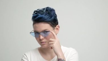 Kısa saçlı, koyu mavi saçlı, beyaz tişörtlü, açık arkaplanda genç bir kız. Bir kadın stüdyoda poz veriyor, gülümsüyor ve duygularını gösteriyor..