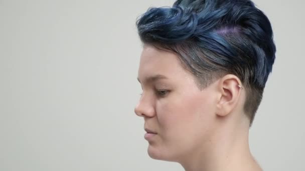 Una joven con un corte de pelo corto de hada y cabello azul oscuro en una camiseta blanca sobre un fondo claro. Una mujer posa en el estudio, sonríe y muestra emociones. — Vídeo de stock