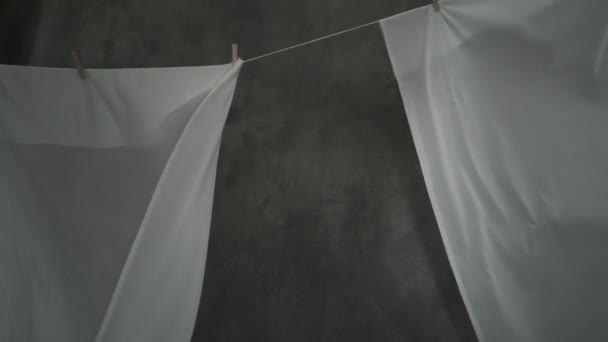 Weiße Stoffbahnen werden auf Wäscheklammern an einem Seil getrocknet. Das wellige Gewebe wiegt sich im Wind. Abstrakter grauer Hintergrund. — Stockvideo