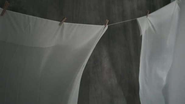 Weiße Stoffbahnen werden auf Wäscheklammern an einem Seil getrocknet. Das wellige Gewebe wiegt sich im Wind. Abstrakter grauer Hintergrund. — Stockvideo
