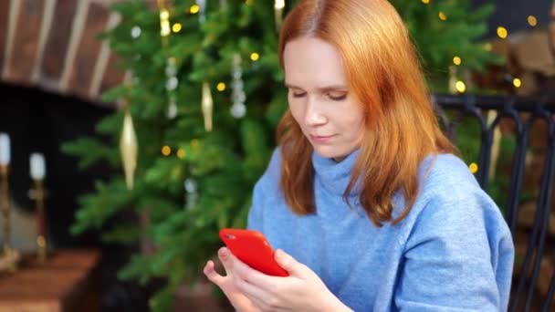 Egy okostelefonos nő ül a karácsonyfánál. közösségi hálózatok — Stock videók