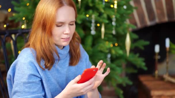 Uma mulher com um smartphone nas mãos está sentada na árvore de Natal. redes sociais — Vídeo de Stock