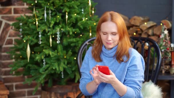 Een vrouw met een smartphone in handen zit aan de kerstboom. sociale netwerken — Stockvideo