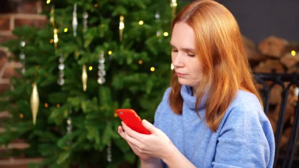 Een vrouw met een smartphone in handen zit aan de kerstboom. sociale netwerken — Stockvideo