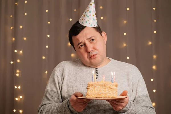 Sad man in festive cap with birthday cake blows out candles.sadness due to aging — Stock Photo, Image