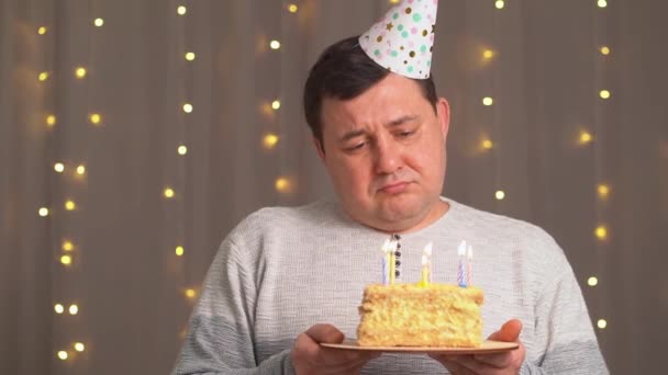 Hombre triste en gorra festiva con pastel de cumpleaños sopla candles.sadness debido al envejecimiento — Vídeos de Stock