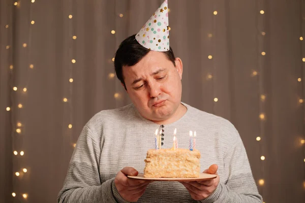 Sad man in festive cap with birthday cake blows out candles.sadness due to aging — Stock Photo, Image