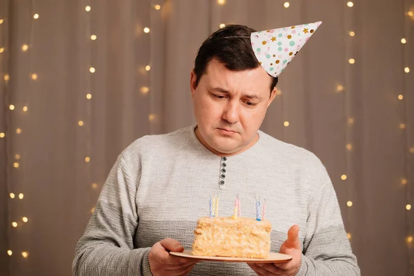 Sad man in festive cap with birthday cake blows out candles.sadness due to aging — Stock Photo, Image