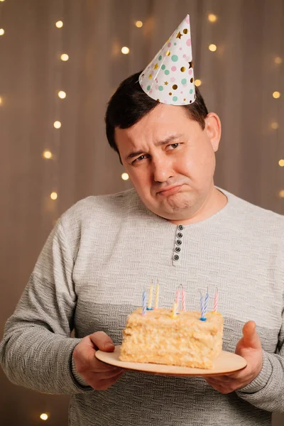 Sad man in festive cap with birthday cake blows out candles.sadness due to aging — Stock Photo, Image