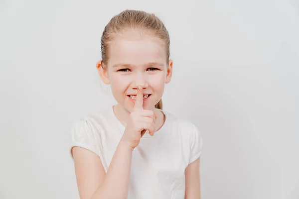 Little girl with in a white T-shirt holds index finger in front of lips. — Stock Photo, Image