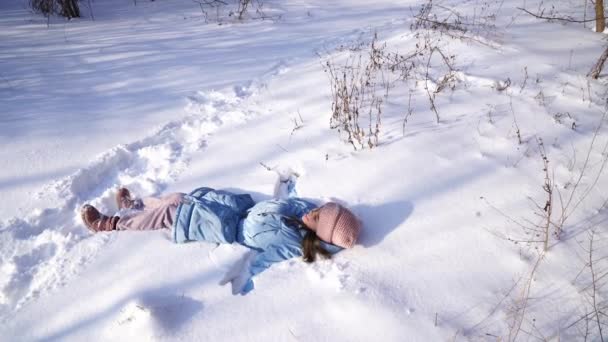 Tonåring flicka i blå rock och rosa mössa faller i snön och gör en snö ängel — Stockvideo