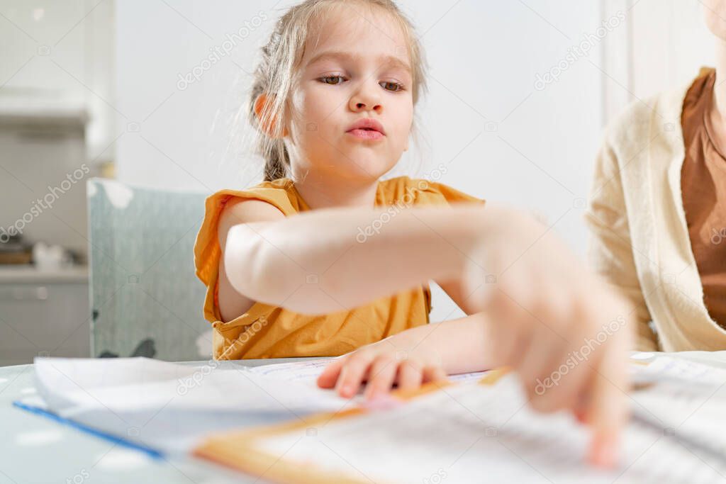 A little schoolgirl does her homework on the textbook. mom sits next to her