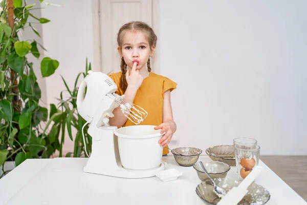 Niña pequeña prepara masa o crema en un mezclador sumergible y lame sus dedos —  Fotos de Stock