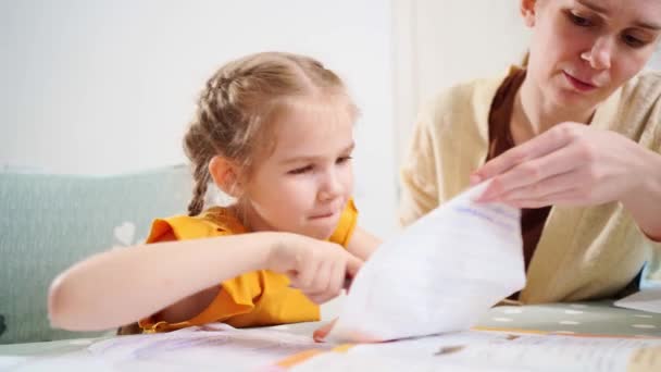 Een schoolmeisje doet haar huiswerk. moeder zit naast haar legt uit en helpt — Stockvideo