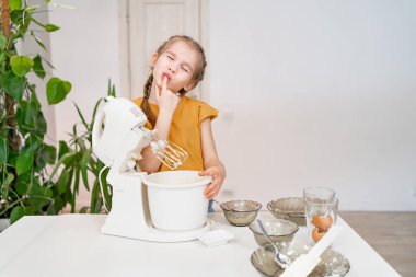 Küçük kız hamuru denizaltı mikserinde hazırlıyor ve parmaklarını yalıyor. Modern ev aletleri çocuklar için bile uygun ve basittir. Yemek pişirme hobisi. tatil.