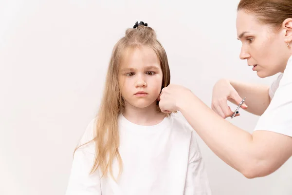A woman does haircut a funny girl's on a white background. Mom cuts hair her daughter at home. The child is afraid and regrets the severed hair.