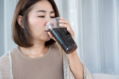 Asyalı orta yaşlı kadın evde plastik bardaktan tatlı soda ya da kola içiyor. Sağlıksız beslenme, şeker bağımlılığı, ferahlatıcı içecek ve yetişkinlerde diyabet veya obezite riski kavramı.