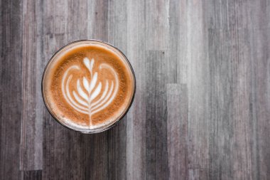 Sütlü kahve. Çekici desenli. Ahşap bir masada cam bir fincanda.