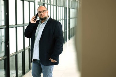 Sırt çantalı orta yaşlı bir adam şık, cam panelli ofis koridorunda dikilirken akıllı telefonunu kontrol ederken görülüyor..