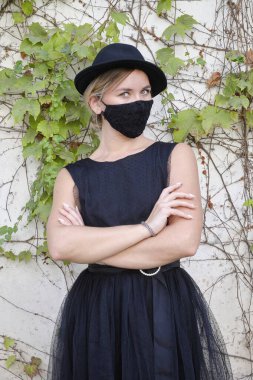 Siyah şapkalı, siyah elbiseli ve COVID maskeli genç kız.