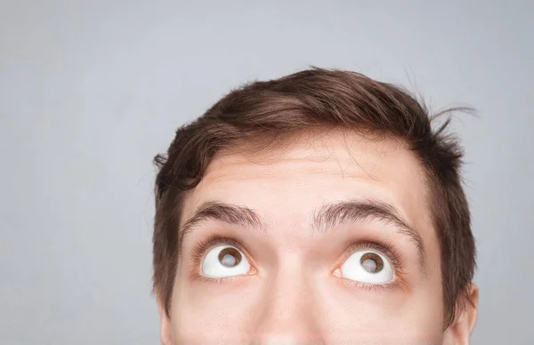 Man\'s eyes looking up on the light background. Empty place for a text or object. Close-up shot of shocked young man with round eyes, top half head. Eyes looking on top