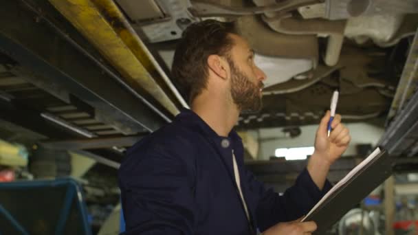 Mechanic inspecteert de auto onderstel manier en maakt een opmerking van zijn inspectie — Stockvideo