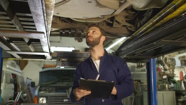 Mechanic inspecteert de auto onderstel manier en neemt een nota over zijn inspectie — Stockvideo