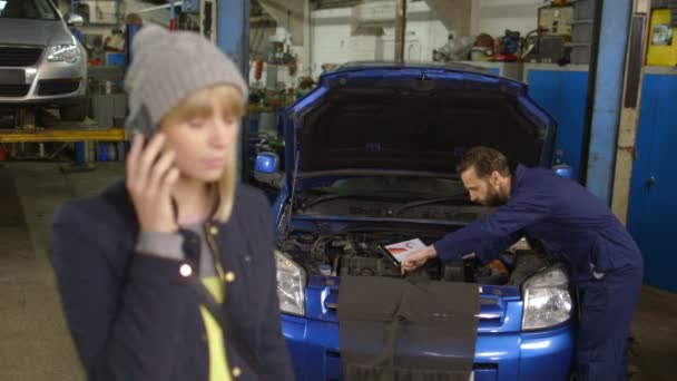 Cliente feliz en el teléfono, mientras que un mecánico trabaja en un coche en el fondo — Vídeos de Stock