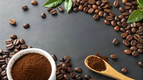 Koyu dokulu bir yüzey ustalıkla kavrulmuş kahve çekirdekleri öğütülmüş kahve bardağı ve içinde kahve tozu bulunan tahta bir kaşık ile düzenlenir.