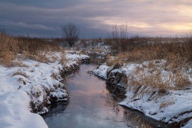 Gün batımında karlı bir tarlada akan dere.