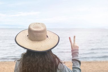 Şapka takan, barış yapan, parmaklarıyla sevişen, denize bakan bir kız. Turizm ve tatil kavramı.