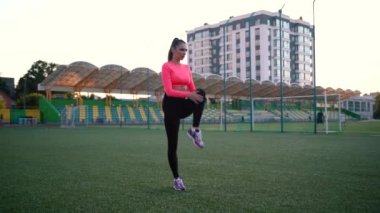 Spor giyim sektöründe spor giyinen kadın stadyumdaki yeşil futbol sahasında, beyaz işaretli spor minderleri üzerinde çeşitli egzersizler yapıyor. Açık hava sporları. Online eğitim. Sıcak yaz günü. sağlıklı yaşam tarzı kavramı