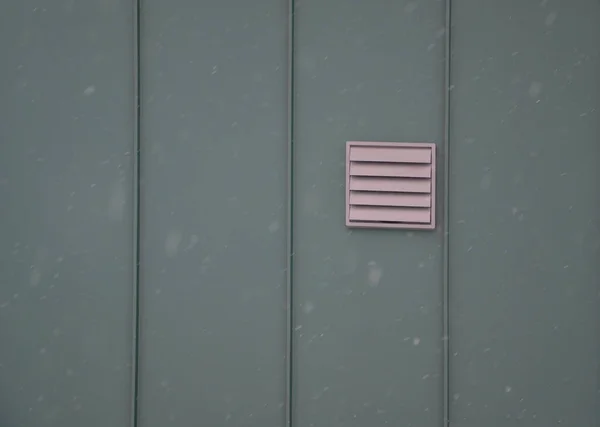 lattice with lamella opening on the wall of a green sheet metal building. a square grille with blinds protects the fan from snow. opens when ventilation starts