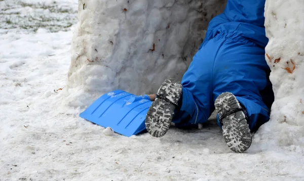 一个穿着冬装的小孩穿着蓝色塑料铲子在雪地上走着 将建造一个厕所 小男孩爬到冰洞里 房子里 他有手套和一把有木制把手的铲子 — 图库照片