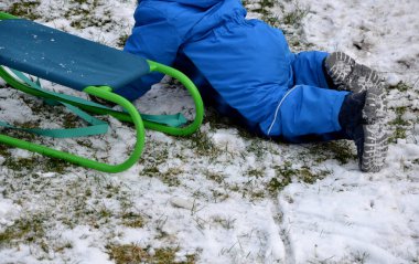 Kış tulumu giymiş küçük bir çocuk kızağını çekmeye çalışıyor ama yine de dizlerinin üzerinde kar kalıntılarının üzerinde sürünüyor. Bir ip tutuyor ve metalik yeşil bir koltukta oturuyor, bahçede kızakla kayıyor.