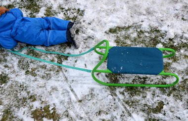 Kış tulumu giymiş küçük bir çocuk kızağını çekmeye çalışıyor ama yine de dizlerinin üzerinde kar kalıntılarının üzerinde sürünüyor. Bir ip tutuyor ve metalik yeşil bir koltukta oturuyor, bahçede kızakla kayıyor.