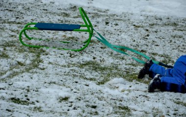 Kış tulumu giymiş küçük bir çocuk kızağını çekmeye çalışıyor ama yine de dizlerinin üzerinde kar kalıntılarının üzerinde sürünüyor. Bir ip tutuyor ve metalik yeşil bir koltukta oturuyor, bahçede kızakla kayıyor.