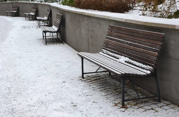 snowy benches near the supporting concrete gray wall in the park. Paving and metal low fences protect ornamental flower beds from dog urine