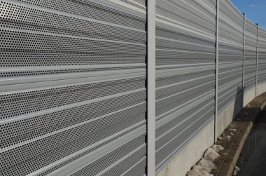 Metal delikli metal levhalardan oluşan ses bariyeri. Gri ve gümüşi koruyucu çitler gürültülü bir yoldan evleri ayırıyor. Gözenekli madde lastik sesini iyi parçalar.