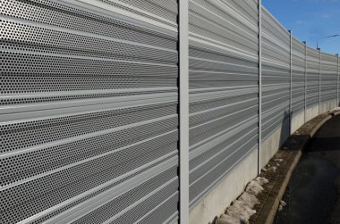 Metal delikli metal levhalardan oluşan ses bariyeri. Gri ve gümüşi koruyucu çitler gürültülü bir yoldan evleri ayırıyor. Gözenekli madde lastik sesini iyi parçalar.